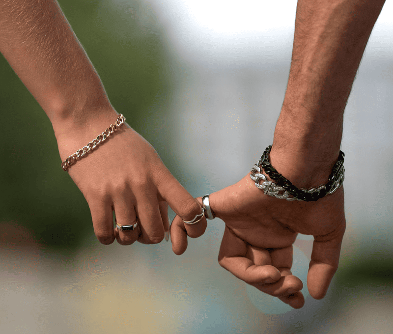 Two people holding hands outdoors, wearing gold and black chain bracelets and rings.