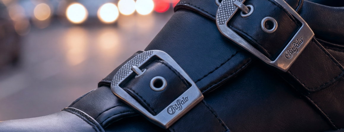 Close-up of black leather boots with silver metal buckles, featuring engraved brand details, set against a blurred city street background with evening lights.
