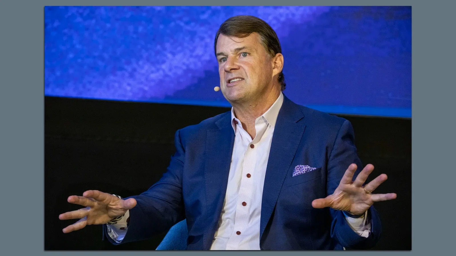 Ford CEO Jim Farley, wearing a white collared shirt and a blue blazer, spreads his hands while speaking onstage at a conference. 