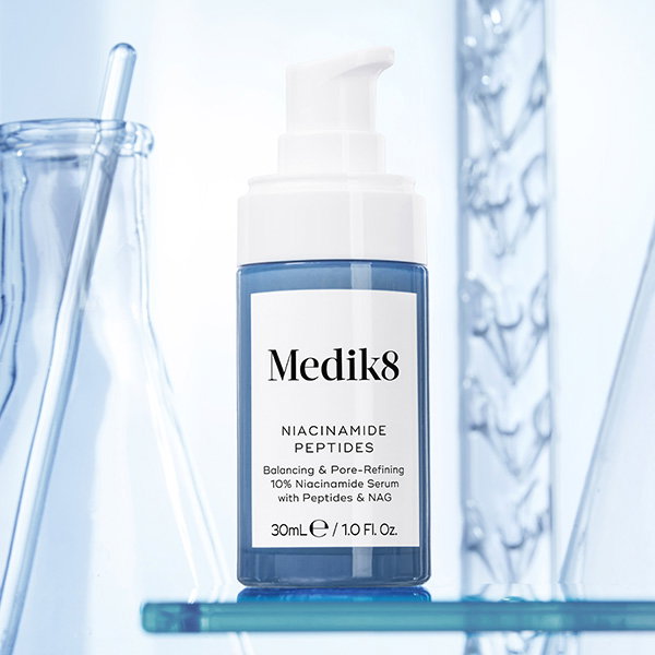 The Medik8 Niacinamide Peptides bottle is centered on a glass shelf. Behind the bottle, blurred laboratory glassware, including beakers and a graduated cylinder, are visible against a bright blue and white background, emphasizing a clinical or scientific aesthetic.
