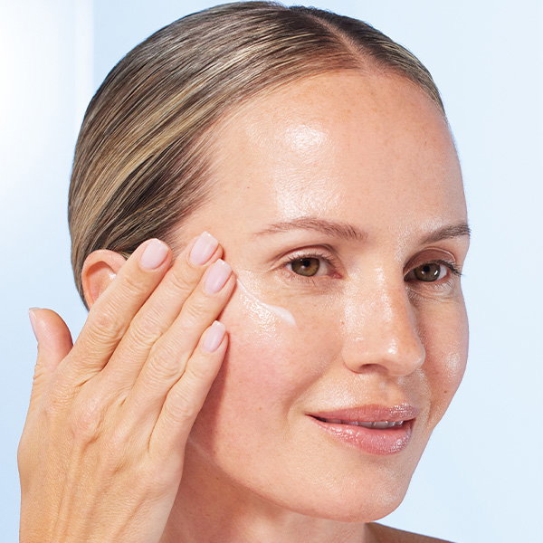 A close-up photograph of a person with light-brown hair pulled back into a sleek bun. They are smiling and applying a white serum to their cheek with their fingertips, which feature a light pink manicure. The background is a soft, blurred light blue.