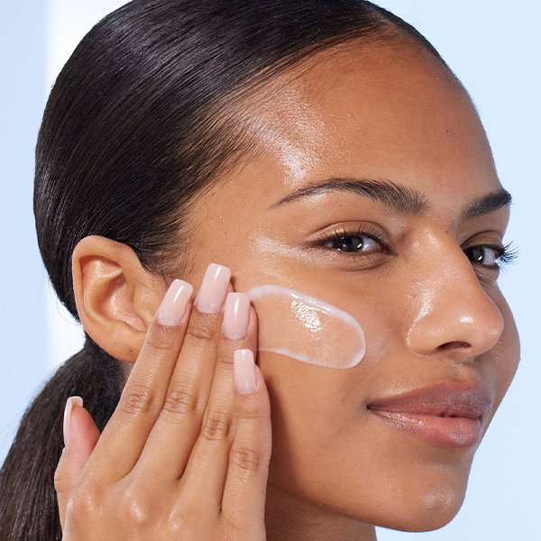 A close-up of a person with dark hair in a ponytail applying a thick streak of creamy white serum across their cheek. They are using their hand, which has long, square-tipped pale pink nails, to blend the product into their glowing skin.