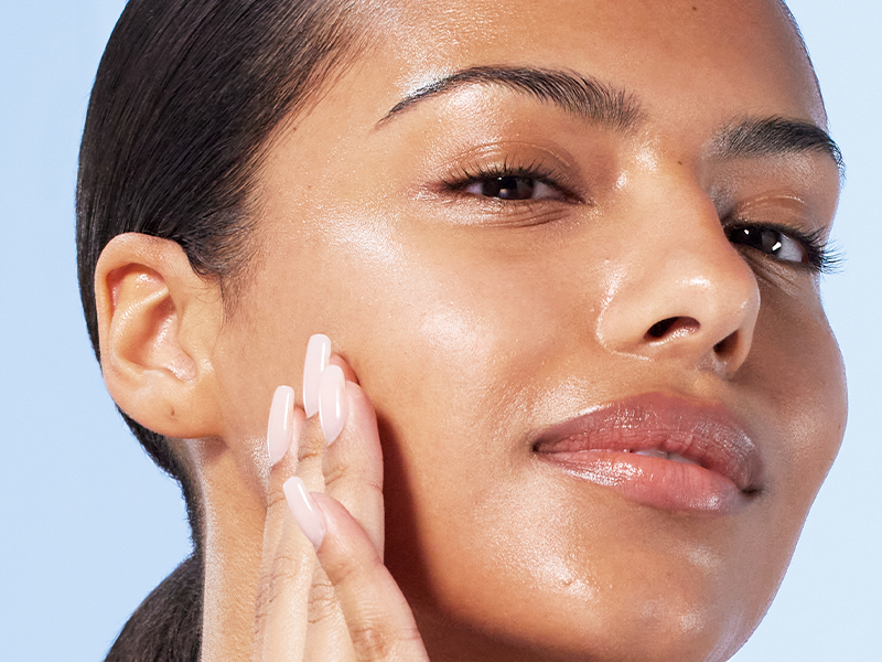 A close-up photograph of a person with glowing, hydrated skin and dark hair pulled back. They are lightly touching their cheek with their fingers, showcasing long, pale pink manicured nails. The background is a solid light blue.