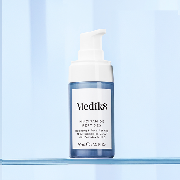 A bottle of Medik8 Niacinamide Peptides serum sits on a reflective blue surface against a light blue, vertically striped background. The serum comes in a translucent blue bottle with a white pump dispenser and a white label featuring black text.