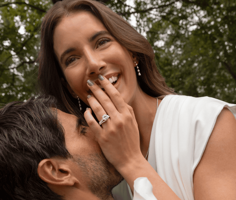 Man and woman embracing outdoors, woman wearing a white gold round-cut diamond engagement ring and diamond drop earrings.