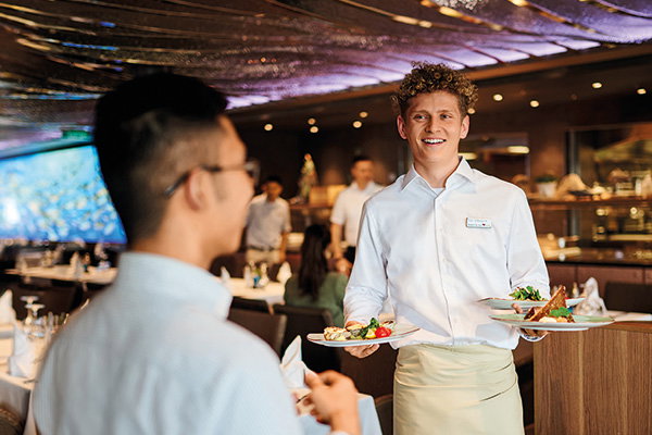In einem Restaurant serviert eine Person in weißer Kleidung zwei Teller mit Speisen an eine andere Person. Im Hintergrund sitzen weitere Gäste.