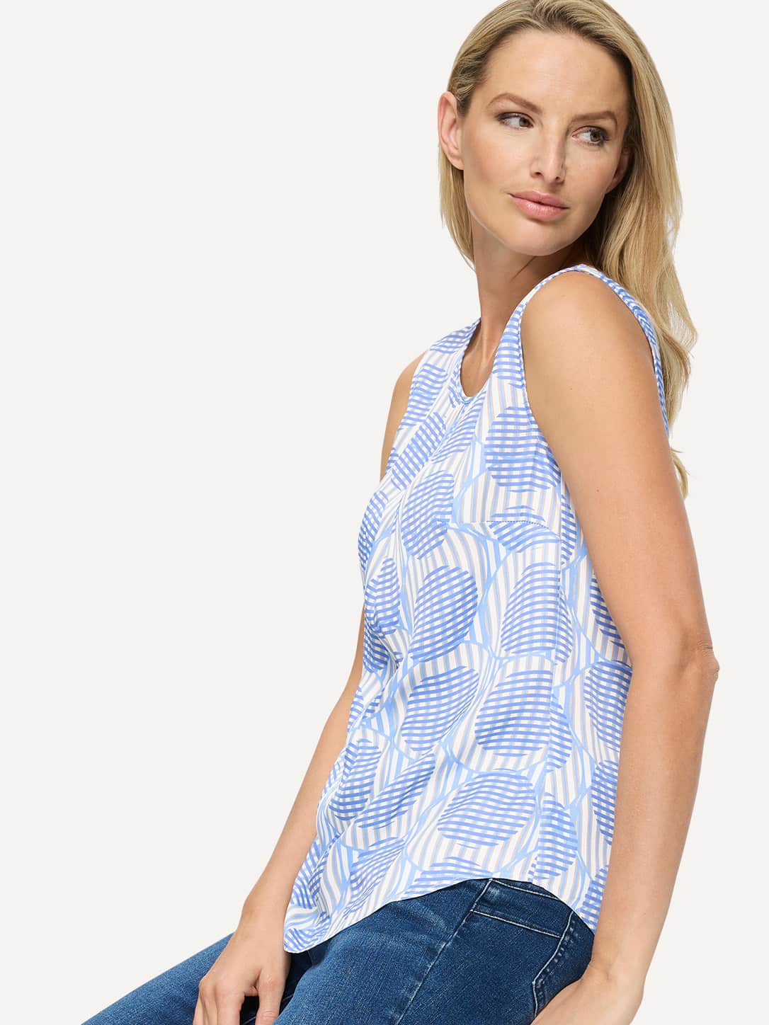 Een model draagt een mouwloos topje met blauw-wit geometrisch patroon, gecombineerd met blauwe jeans, voor een lichte achtergrond. 