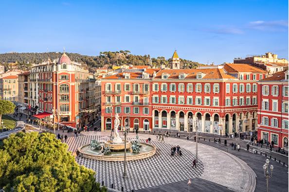 A bustling city square with a fountain, red-roofed buildings, and people walking.
