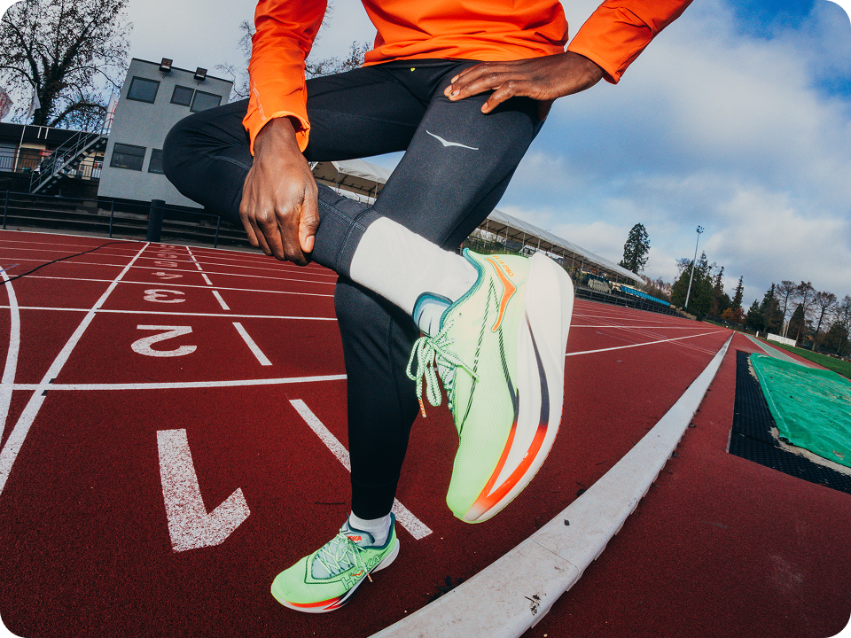An image of an athlete from the waist down, standing on a race track with one leg crossed over the other showing the Cielo X1 3.0 running shoe.