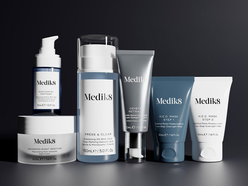 A collection of Medik8 skincare arranged on a sleek dark surface. The lineup includes Crystal Retinal, Advanced Night Restore cream, Niacinamide Peptides, the two-step H.E.O. Mask and Press & Clear exfoliating BHA tonic.
