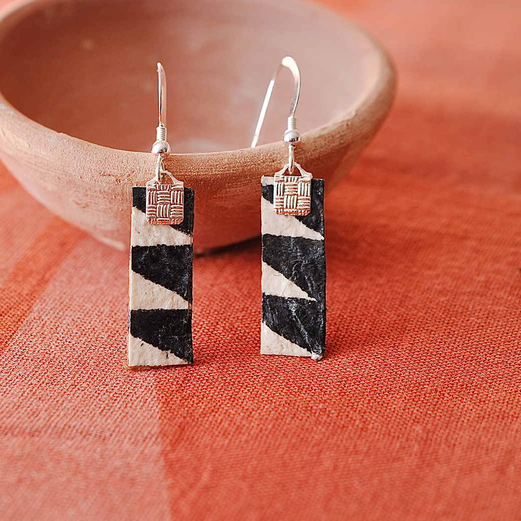 Pair of earrings crafted from Hawaiian kapa with geometric patterns, propped against a terracotta bowl. - Shop Now.