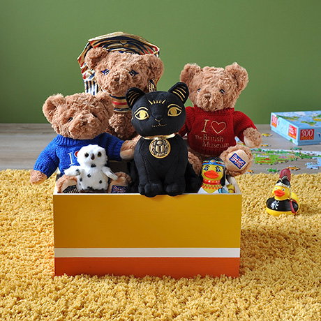 Collection of plush teddies, including Bastet, an Ancient Egypt-inspired teddy, sitting in a toy box on a yellow carpet with a green background. - Shop Children Collection.