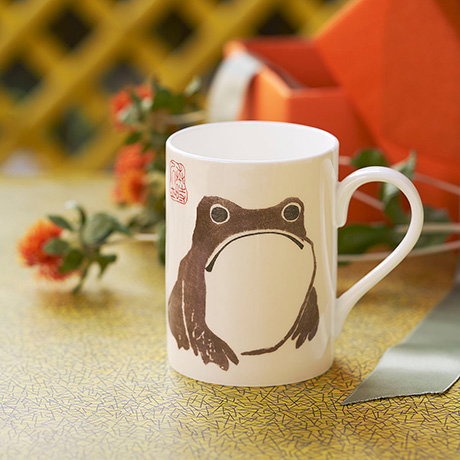 Bone china mug featuring the grumpy-looking frog by Matsumoto Hōji, with orange flowers and an open gift in the background. - Shop Gifts Collection. 