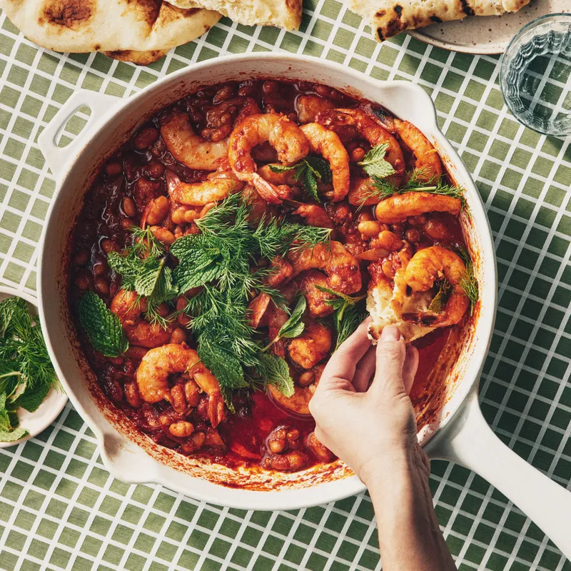 A healthy dinner of shrimp and white beans in a skillet with tomato sauce, topped with dill and mint, and served with toasted naan.