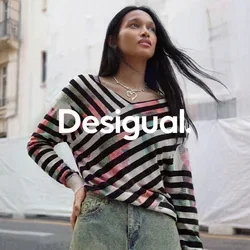 Femme aux longs cheveux foncés portant un pull rayé multicolore et une jupe en jean, debout dans une rue de la ville avec des bâtiments historiques.