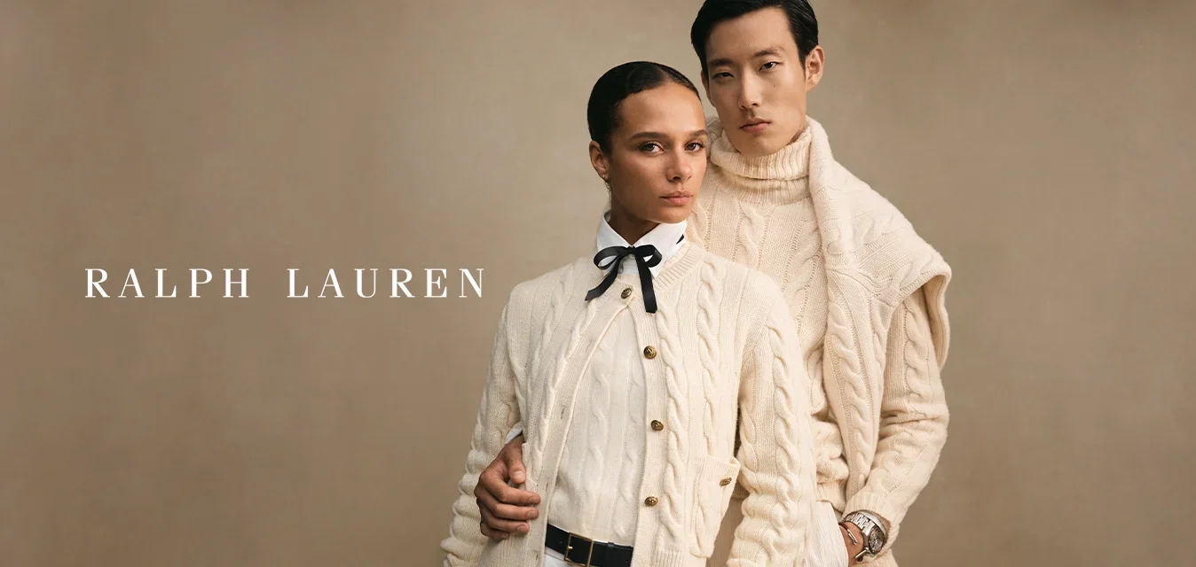 Deux mannequins portant des pulls en tricot torsadé crème posent côte à côte sur un fond beige, l'un arborant un nœud papillon et l'autre une montre.