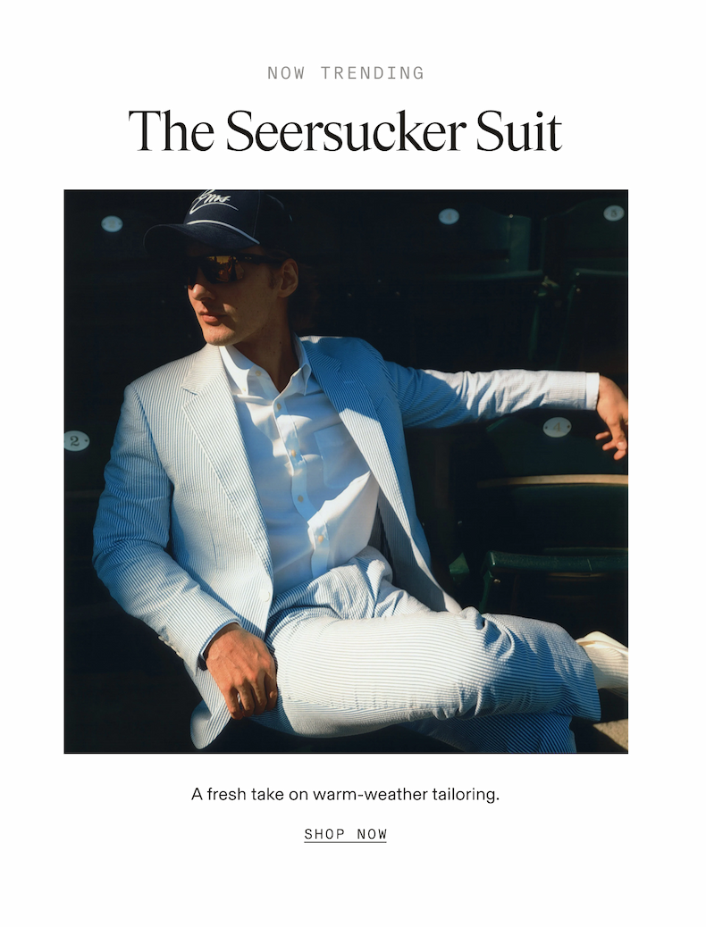 Text-only header with subheading reading: "NOW TRENDING" followed by "The Seersucker Suit." Below is a lifestyle photo of a man seated in stadium chairs, wearing a light blue and white seersucker suit with a white dress shirt, sunglasses, and a navy baseball cap. Text below the photo reads: "A fresh take on warm-weather tailoring. SHOP NOW"
