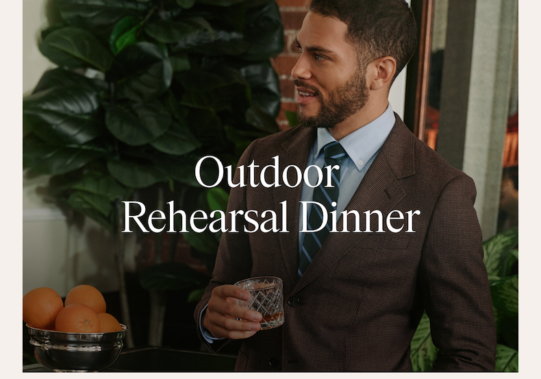 A man wearing a brown houndstooth suit with a light blue dress shirt and navy striped tie, holding a glass of whiskey and smiling. Lush green foliage visible in the background. Text overlay reads: "Outdoor Rehearsal Dinner"