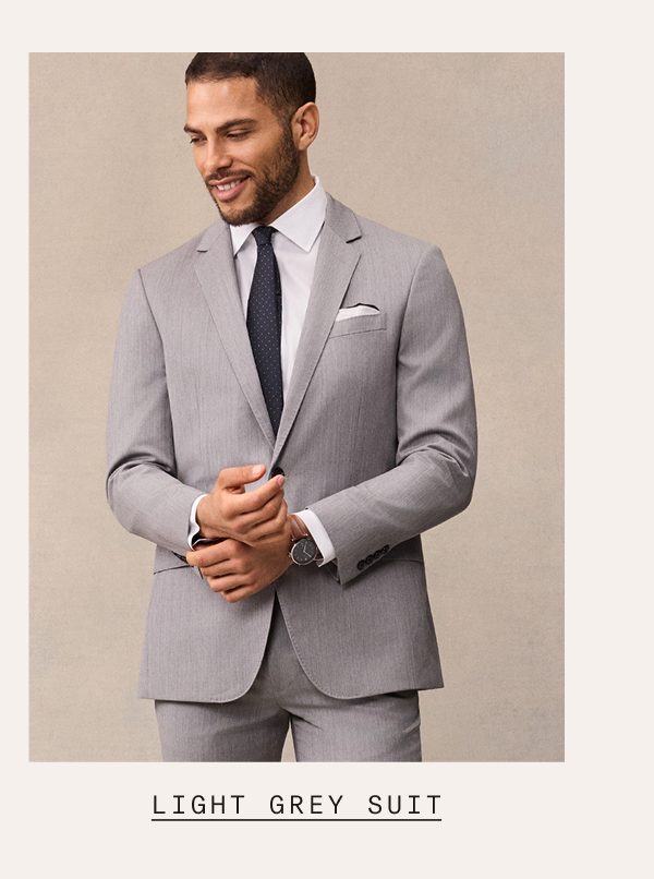A man wearing a light grey suit with a white dress shirt, navy tie, and white pocket square, adjusting his cuffs and smiling. Text below reads: "LIGHT GREY SUIT"