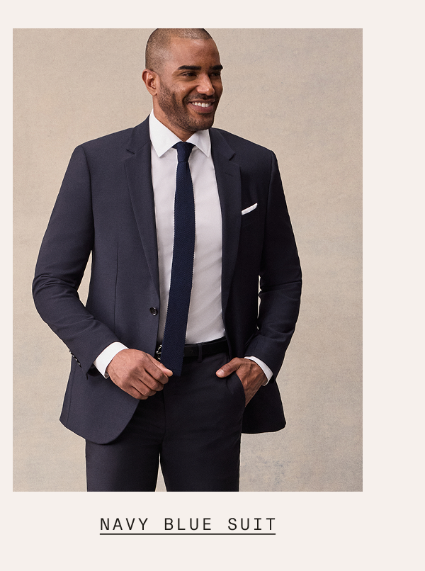 A man wearing a navy blue suit with a white dress shirt, navy knit tie, and white pocket square, smiling with one hand in his pocket. Text below reads: "NAVY BLUE SUIT"