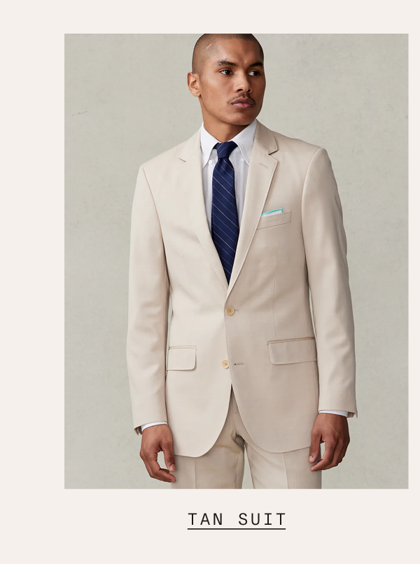 A man wearing a tan suit with a white dress shirt, navy striped tie, and mint pocket square, standing against a light grey background. Text below reads: "TAN SUIT"