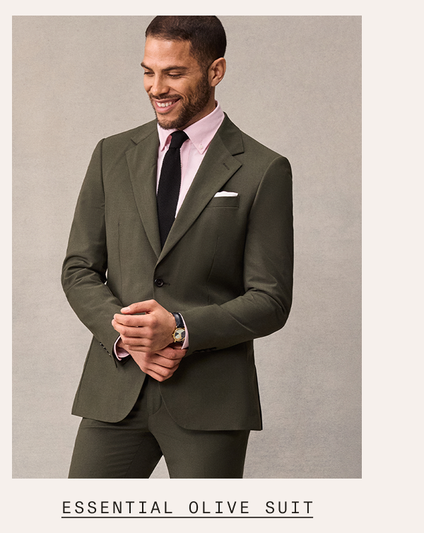 A man wearing an essential olive suit with a pink dress shirt, black tie, and white pocket square, smiling and adjusting his cuffs. Text below reads: "ESSENTIAL OLIVE SUIT"