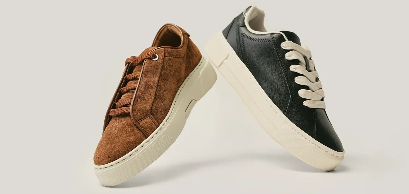 Par de zapatillas informales bajas, una de ante marrón y otra de cuero negro, ambas con suelas blancas y cordones a juego, exhibidas sobre un fondo claro.