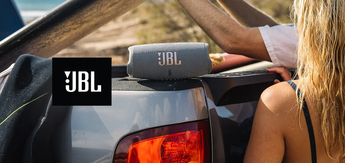 Altavoces portátiles JBL grises sobre la caja de una camioneta, flanqueados por un traje de neopreno negro y una camiseta de manga larga blanca. Tabla de surf sostenida en alto, ambiente de playa.