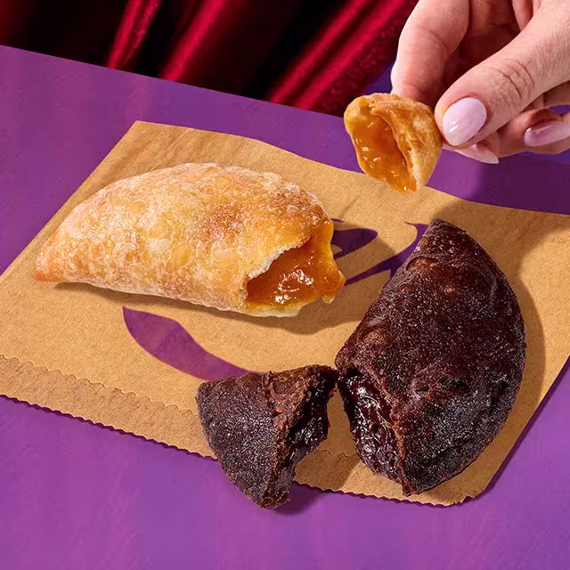 A hand holding a piece of a Caramel Empanada next to a Chocolate Fudge & Caramel Empanada on purple table.