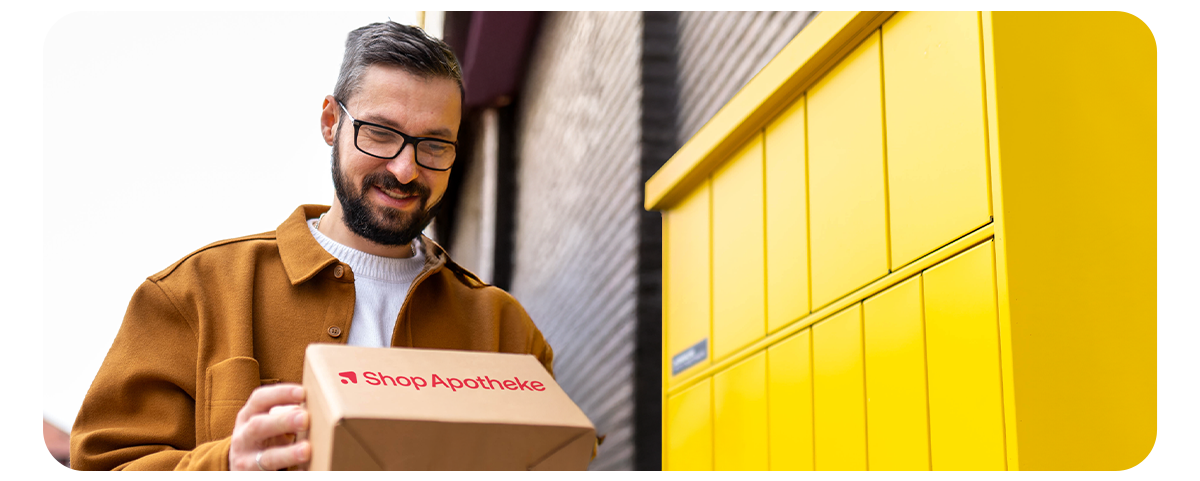 Mann holt ein Paket von Shop Apotheke an einer gelben Packstation ab. Text: „Lieferung an die Packstation – ganz ohne Postnummer. Einfach im Check-out auswählen & flexibel abholen.“ Button: „Jetzt entdecken“.