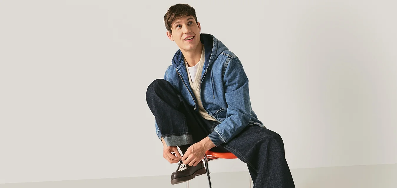 Joven sentado en una silla atándose unos zapatos de cuero marrón oscuro, vestido con chaqueta vaquera, camisa blanca y vaqueros con puños anchos, sobre un fondo liso.