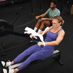 Máquina de remo situada en un gimnasio. Dos usuarios con ropa deportiva; uno con leggings morados y top, y el otro con una camiseta verde. Suelo y equipo de color negro.