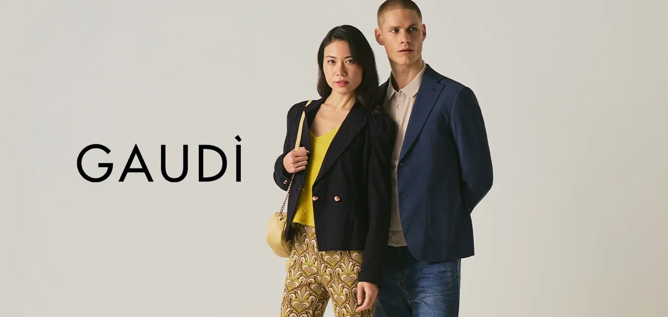 Mujer con pantalones estampados y blazer negro junto a un hombre con blazer azul y vaqueros, sobre un fondo liso.