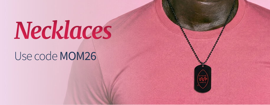 Close-up on the upper chest of a man wearing a pink t-shirt and a MedicAlert necklace. The necklace has a black bead chain and a black, rectangular medical ID tag with the 'MedicAlert' logo in red. The red title text reads 'Necklaces'. Below the title, smaller text says, 'Use code MOM26'.