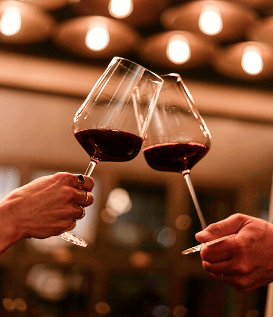 Two glasses of red wine raised in a toast beneath warm ambient lighting.