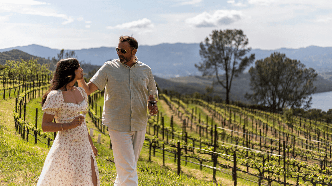 A couple walking through sunlit vineyard rows with rolling hills in the distance.