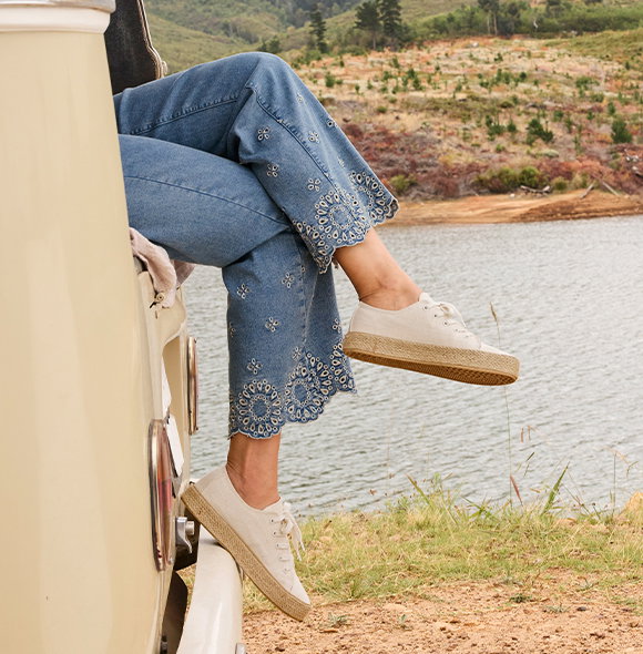Frauenmodel trägt Jeans mit Stickerei, sitzend auf Auto, Beine im Vordergrund überschlagen, Natur und Wasser im Hintergrund.