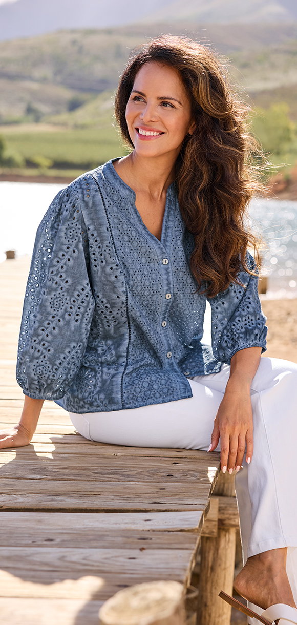 Frauenmodel trägt blaue Bluse mit Muster, weiße Hose, sitzend auf Steg.