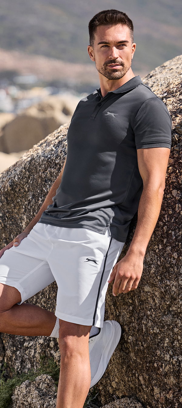 Männermodel trägt weiße Trainingsbermudas und dunkelgraues Funktions-Polo-Shirt, lehnend an Felsen, seitlicher Blick in die Ferne, angewinkeltes Bein.