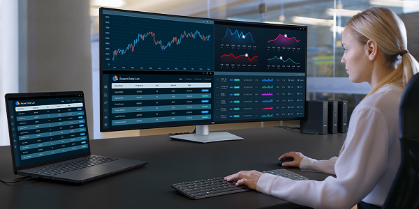An office worker tracking the status of recent orders across multiple dashboards on a widescreen Dell UltraSharp 52 TBT monitor, laptop, wireless keyboard and mouse.