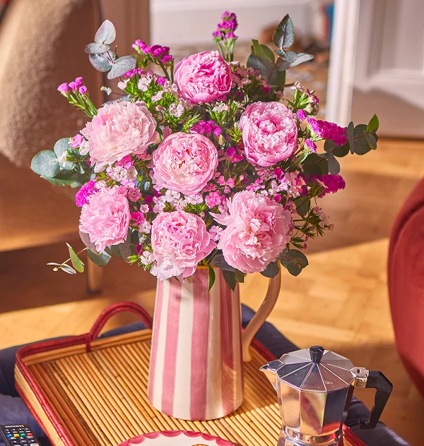 Ein gestreifter Krug präsentiert duftende, pinke Pfingstrosen kombiniert mit farbenfrohem Meerlavendel und frischem Eukalyptus. Das Bouquet steht auf einem Tablett neben einer Espressokanne in einem lichtdurchfluteten Zimmer.