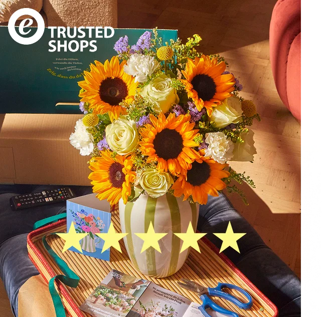 Eine gestreifte Vase mit strahlenden Sonnenblumen , weißen Rosen , Nelken und gelben Trommelstöckchen  steht auf einem Tablett. Das Bild zeigt fünf Sterne und das „Trusted Shops“-Logo. Im Hintergrund ist eine Bloom & Wild Versandbox zu sehen.