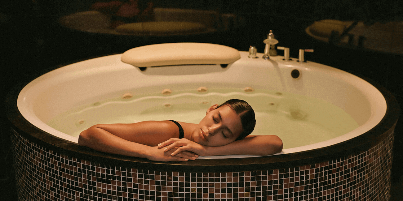 Woman relaxing in a circular spa soaking tub in a dimly lit setting.