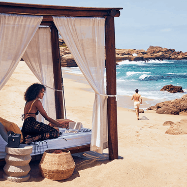 Woman relaxing in a beachfront cabana while a man runs toward the ocean.