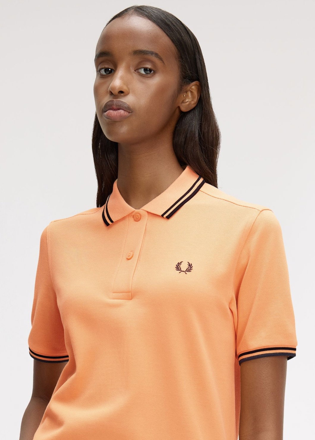 A female model wearing a Fred Perry Shirt in a vibrant peach colour with black twin tipping on the collar
