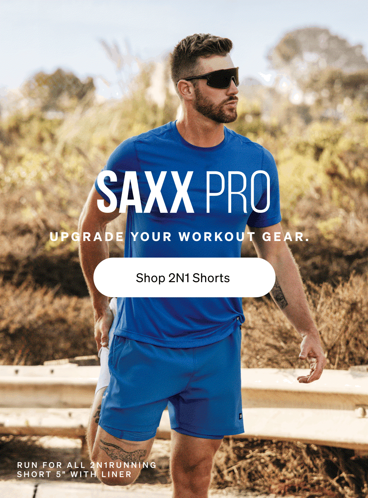 Man in blue athletic shirt and shorts stretching outdoors. Text reads “SAXX Pro. Upgrade your workout gear. Shop 2N1 Shorts.”