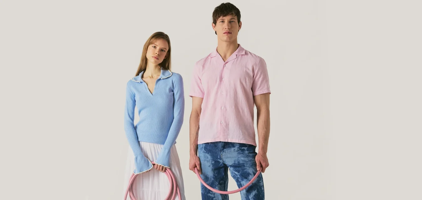 Femme en pull bleu et jupe blanche et homme en chemise rose et jean bleu tenant un tuyau rose enroulé, debout sur un fond clair.