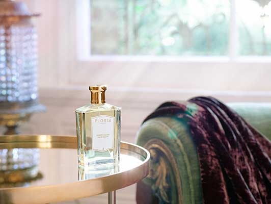 A bottle of Floris White Rose perfume sits on a round gold table next to a green upholstered chair draped with a dark velvet fabric, with sunlight filtering through a window.