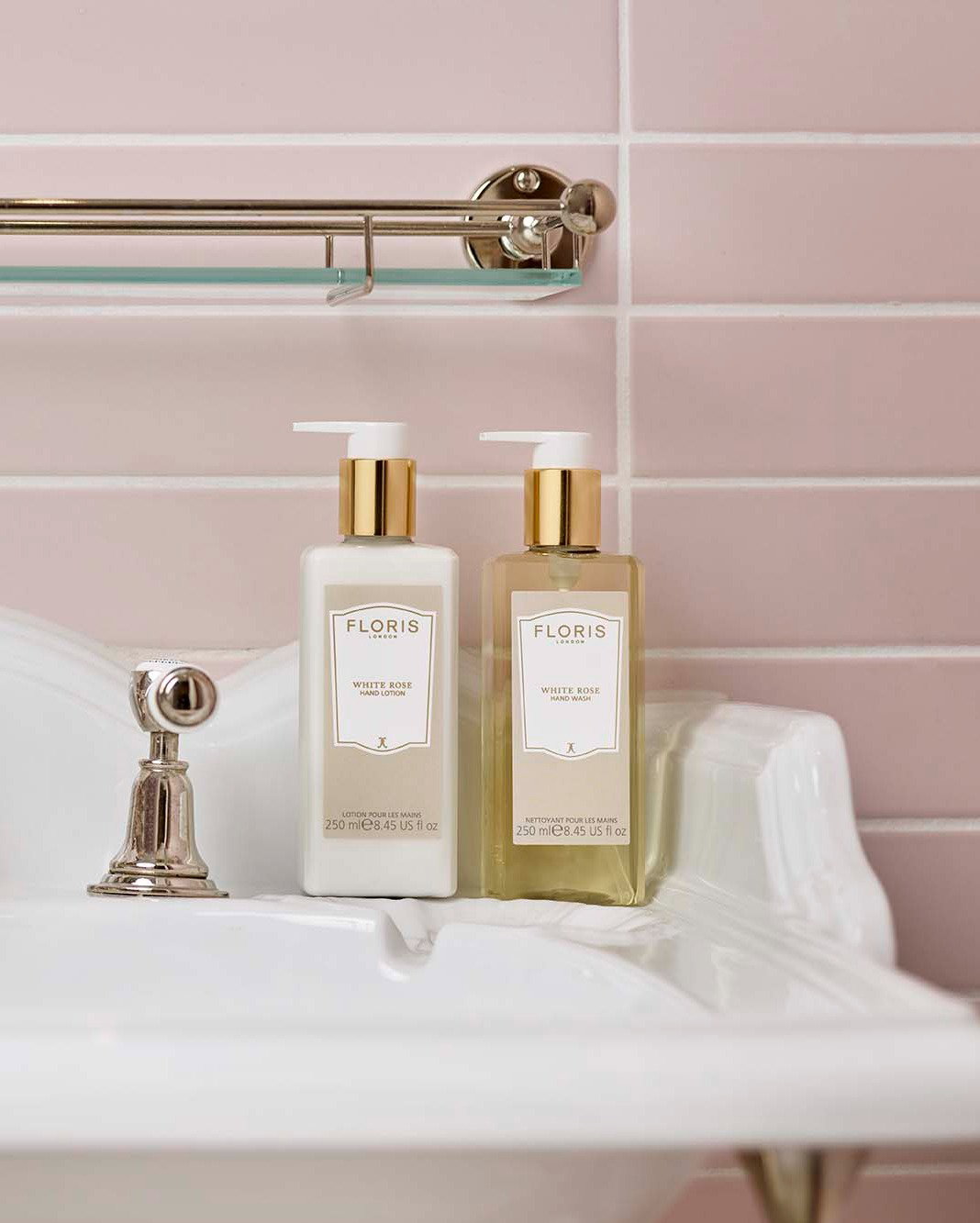 Two bottles of Floris White Rose hand wash and lotion are placed on the edge of a white sink against a wall of pale pink tiles with a glass shelf above.
