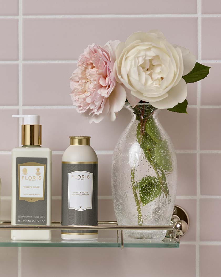 A glass shelf holds two bottles of Floris White Rose toiletries and a clear vase with pink and white flowers, set against a light pink tiled wall.